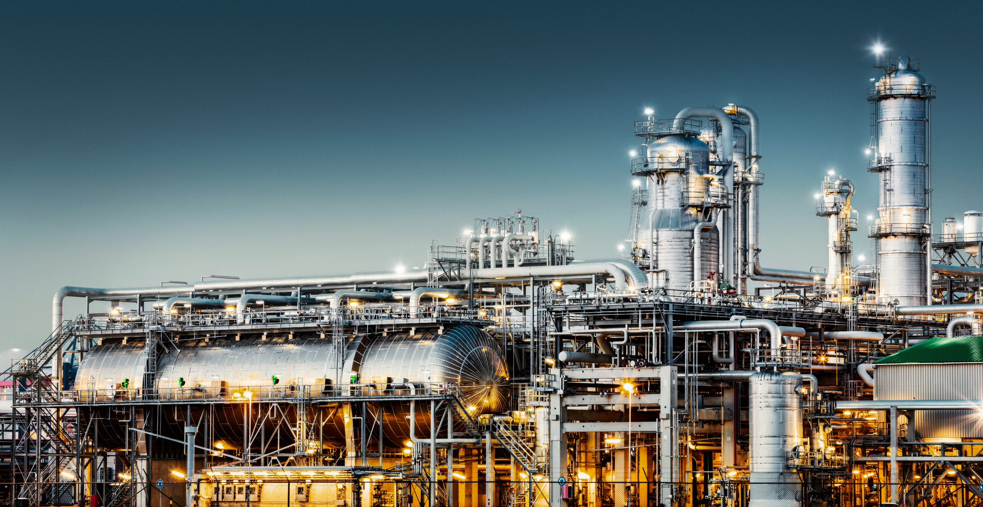 A modern, illuminated refinery is visible at dusk. Lights from the buildings and streets glow. Several distillation towers can be seen as well as glowing moon. Long exposure with tripod.
